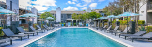 Exterior Canvas apartments pool area, long lap pool, rows of tanning chairs, blue and white umbrellas, residential buildings around pool area, white and blue buildings, lush foliage, photo taken on a sunny day.