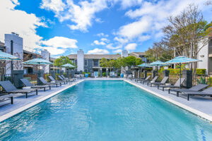 Exterior Canvas apartments pool area, long lap pool, rows of tanning chairs, blue and white umbrellas, residential buildings around pool area, white and blue buildings, lush foliage, photo taken on a sunny day.